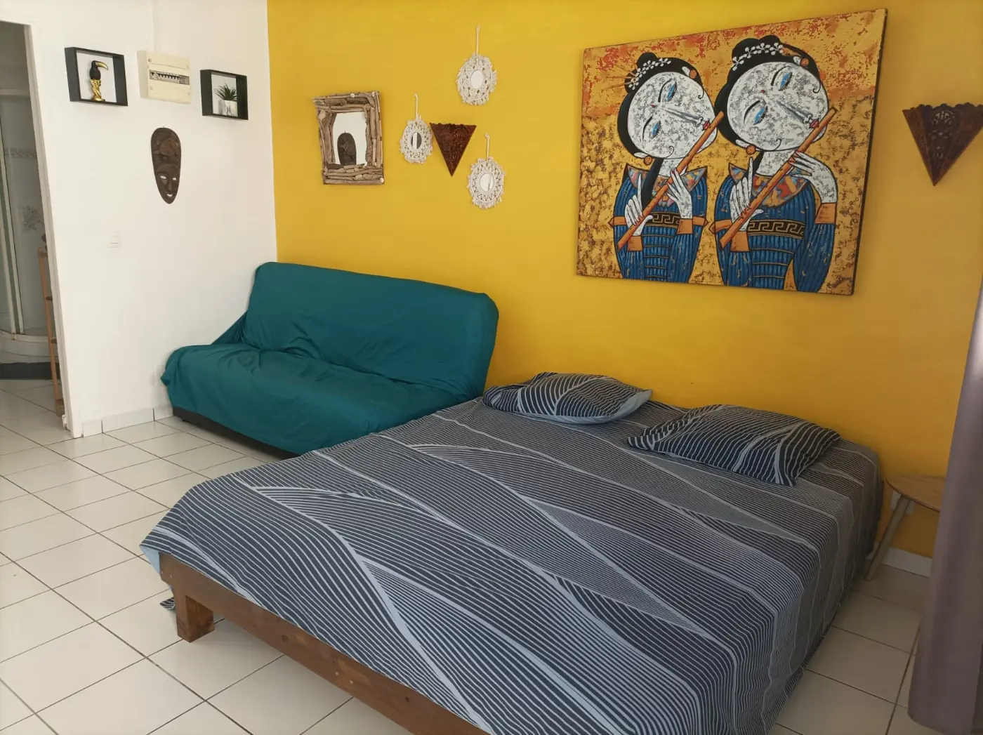 Pièce principale du studio avec lit double, canapé-lit vert et mur jaune orné d'un grand tableau